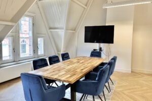 Vergaderruimte aan de Paulus Potterstraat met houten tafel, blauwe stoelen en flatscreen tv aan de muur.